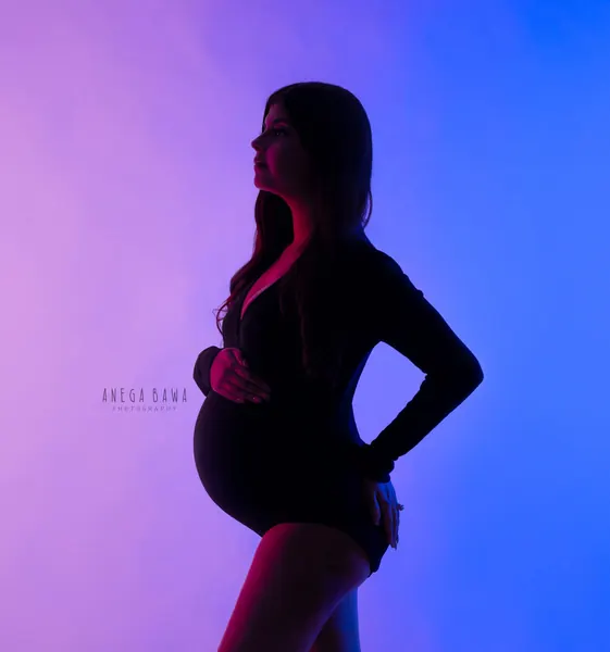 Silhouette of expecting mother in a black long-sleeve bodysuit with a hand on her hip, lit with pink and blue gels against a purple and