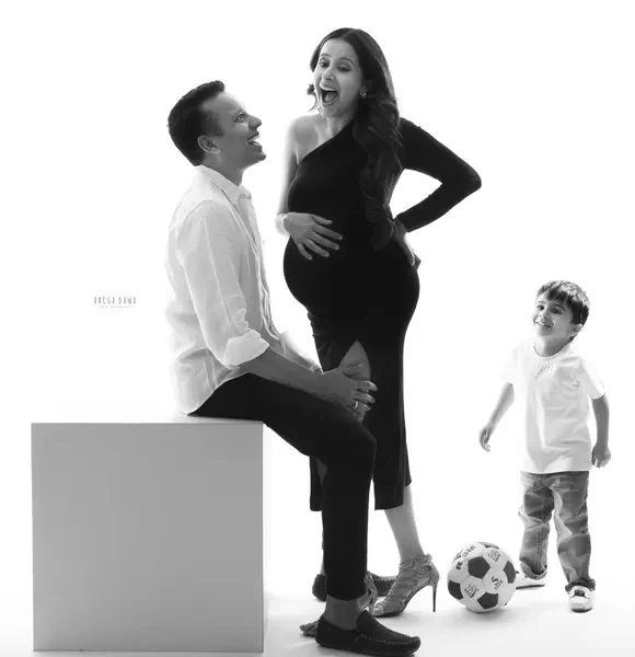 High-key black and white family portrait: husband in a white shirt seated on a white block, wife in a black one-shoulder dress with a slit