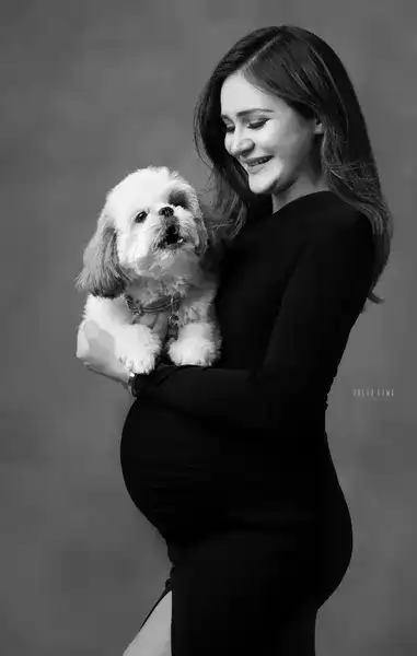 Black and white portrait of smiling expecting mother in a black long-sleeve ruched dress with a high slit holding a small fluffy dog, grey