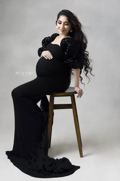 Expecting mother seated on a wooden stool in a black fitted gown with sequin puff sleeves and a long train, looking down at her bump, light