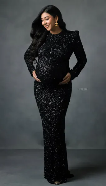 Smiling expecting mother in a fitted black sequin long-sleeve gown with structured shoulders and gold statement earrings, hands on her