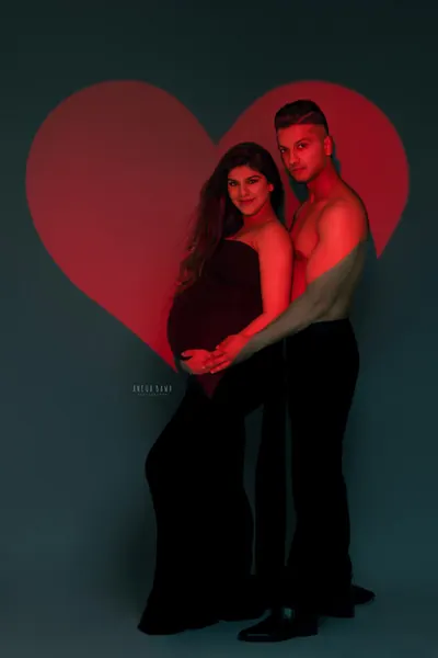 Couple in front of a large glowing red heart, shirtless husband in black trousers holding his wife from behind, wife in a black strapless