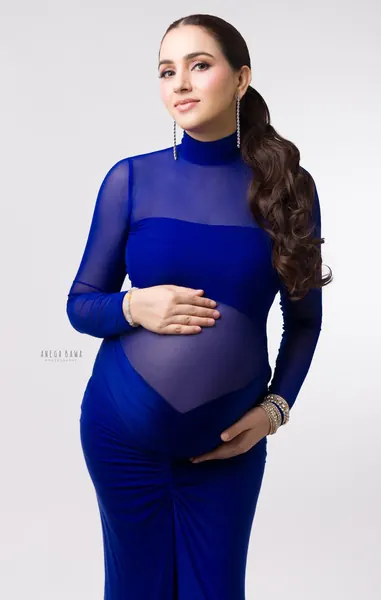 Expecting mother in a cobalt blue gown with sheer mesh panels, high neck and ruched skirt, long drop earrings and ponytail, hands on her