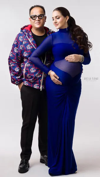 Couple standing side by side, wife in a fitted royal blue sheer-panel long-sleeve gown with a high ponytail, husband in a patterned zip
