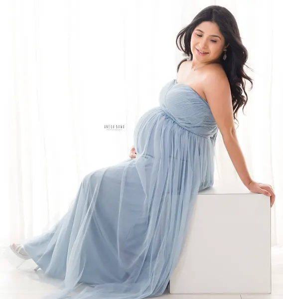 Expecting mother seated on a white block in a dusty blue tulle strapless gown, looking down with a smile, long wavy hair, bright white