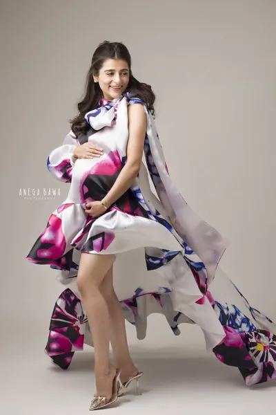 Expecting mother in a floral-print satin mini dress with a flowing cape and train in white, magenta and blue, with clear heels, hand on her