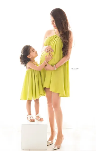 Mother and young daughter in matching lime green flowing mini dresses, daughter standing on a white block touching her mother's bump