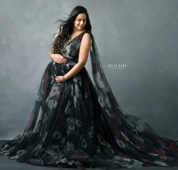 Expecting mother in a V-neck dark floral tulle gown with a sweeping train and a sheer shoulder cape, looking down at her bump, hair