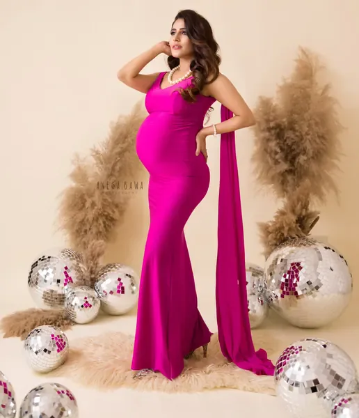 Expecting mother in a fuchsia pink fitted gown with a draped side sash and pearl necklace, posing with silver disco balls and dried pampas