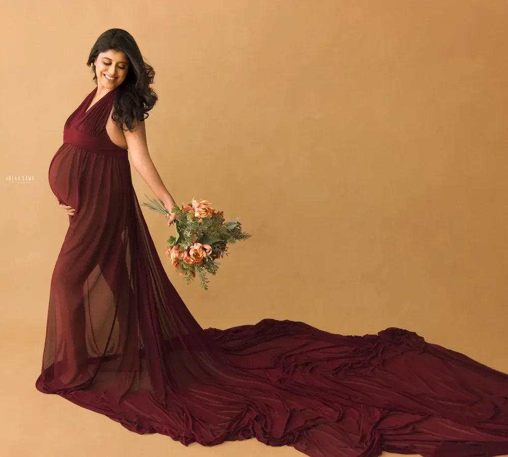 Expecting mother in a maroon halter gown with a sheer maroon train spread out on the floor, holding a small bouquet of peach roses, warm