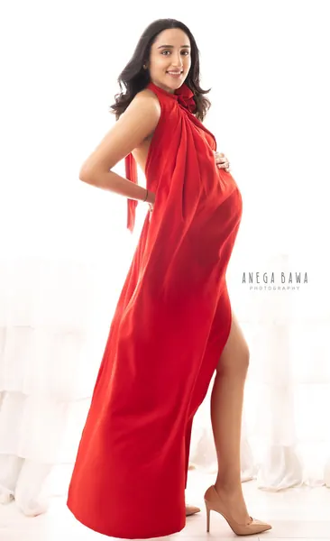 Expecting mother in a red draped halter-style gown with a high slit and nude heels, hand on hip and one on her bump, looking back with a