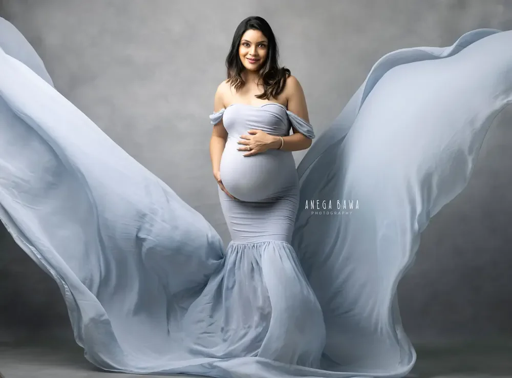 Expecting mother in a dusty blue off-shoulder mermaid gown with huge sheer blue fabric wings flying out on both sides, hands on her bump