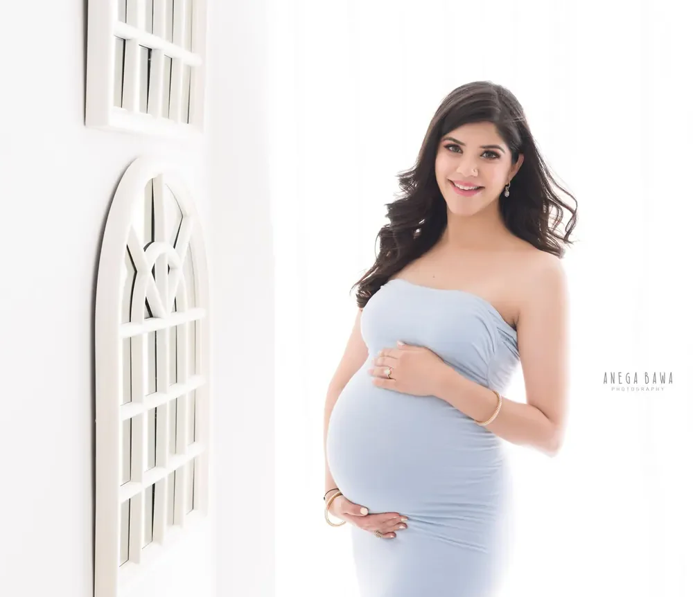 Smiling expecting mother in a pale blue strapless fitted dress with gold bracelets, hands on her bump, white arched window frame in the