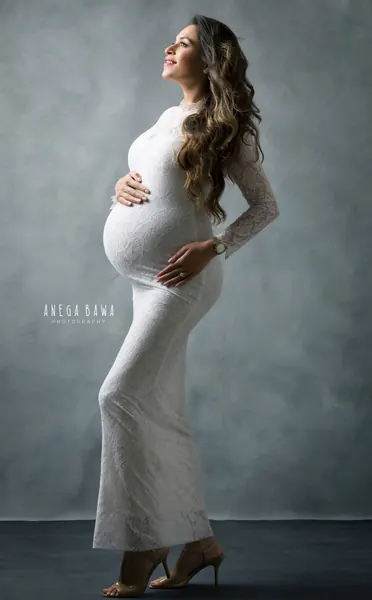 Expecting mother in profile in a fitted white lace long-sleeve gown and nude heels, looking up with a hand on her bump, hair blowing
