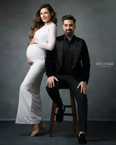 Couple portrait: wife in a white lace long-sleeve fitted gown and heels leaning on her husband, who sits on a wooden stool in a black