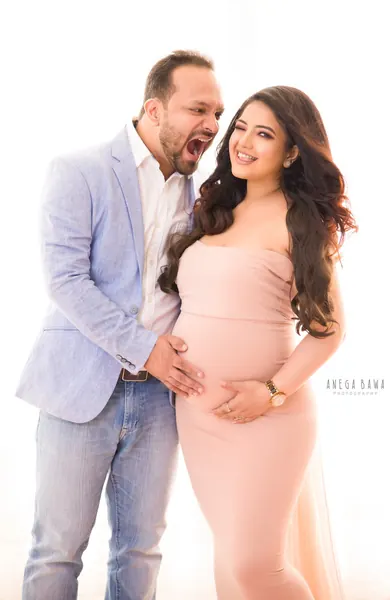 Playful couple, husband in a light blue blazer and jeans pretending to bite his wife's shoulder while holding her bump, wife in a blush