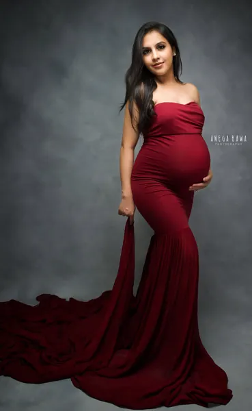 Expecting mother in a deep red strapless mermaid gown with a long train spread on the floor, hand on her bump, looking to the side, grey