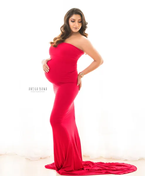Expecting mother in a bright red strapless fitted gown with a spread train, hand on hip and one on her bump, looking down, bright white