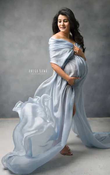 Expecting mother in a pale blue silk drape wrapped off the shoulders with fabric streaming behind her, barefoot, looking down at her bump