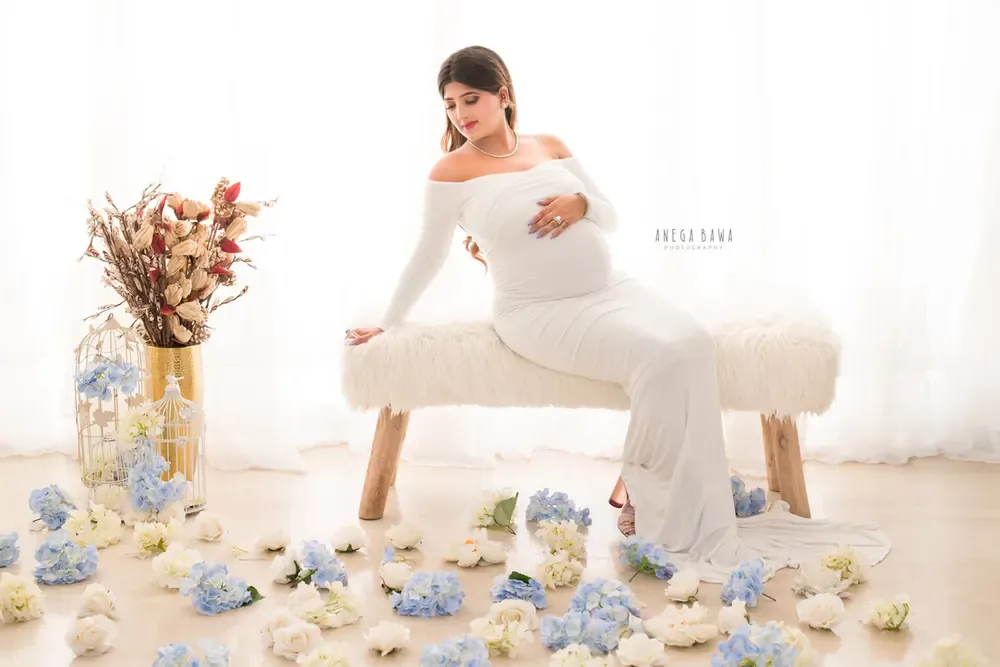 Expecting mother seated on a white fur bench in a white off-shoulder long-sleeve fitted gown, with blue and cream hydrangeas, a birdcage