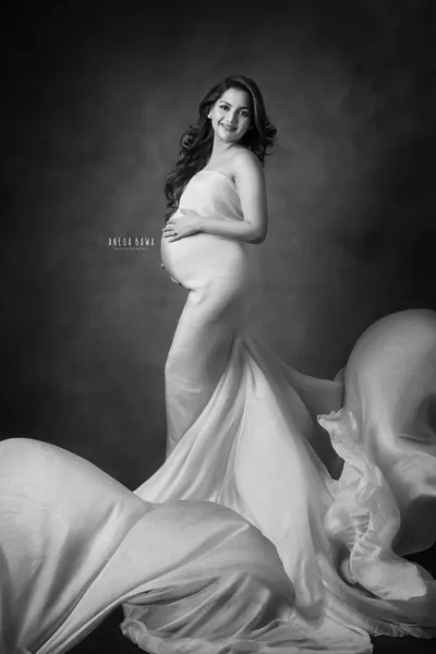 Black and white portrait of smiling expecting mother in a fitted strapless satin gown with large sheer drapes spread at her feet, hand on