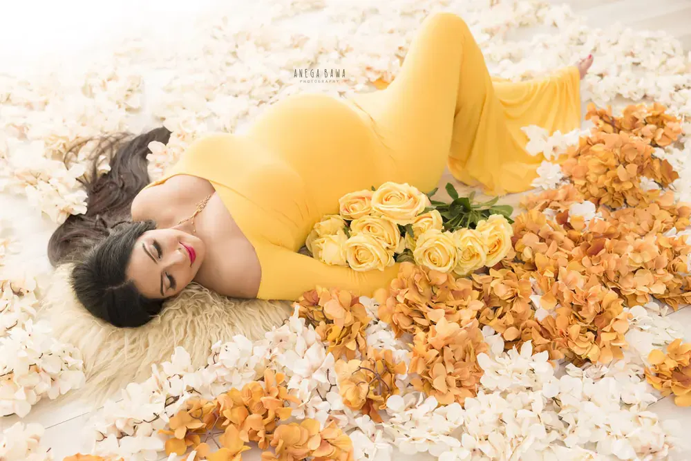 Overhead view of expecting mother lying on a fur rug in a mustard yellow fitted long-sleeve gown among orange and cream hydrangeas, holding