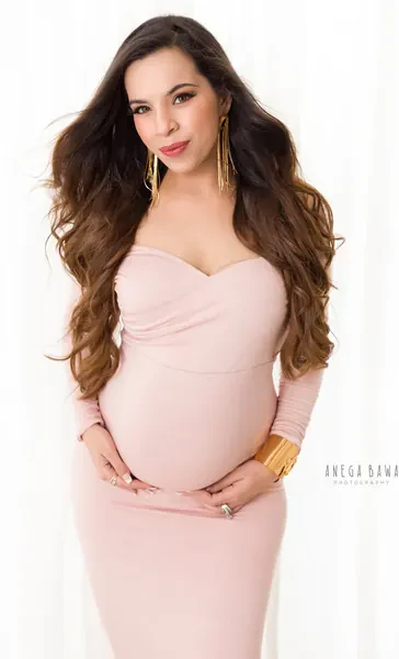 Smiling expecting mother in a blush pink sweetheart-neck long-sleeve fitted dress with long gold drop earrings and a gold cuff, voluminous