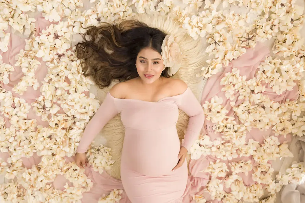 Overhead view of expecting mother lying on a fur rug in a blush pink off-shoulder long-sleeve gown surrounded by cream blossoms, a flower