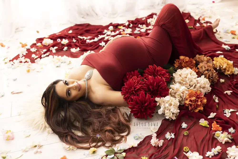 Overhead view of expecting mother lying on a fur rug in a rust red fitted gown, surrounded by dark red and orange flowers and scattered