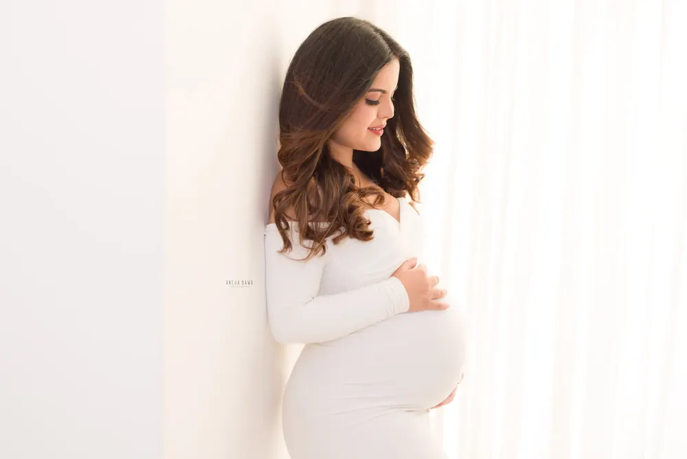 Expecting mother leaning against a white wall in a white off-shoulder long-sleeve fitted gown, looking down at her bump, wavy hair, soft