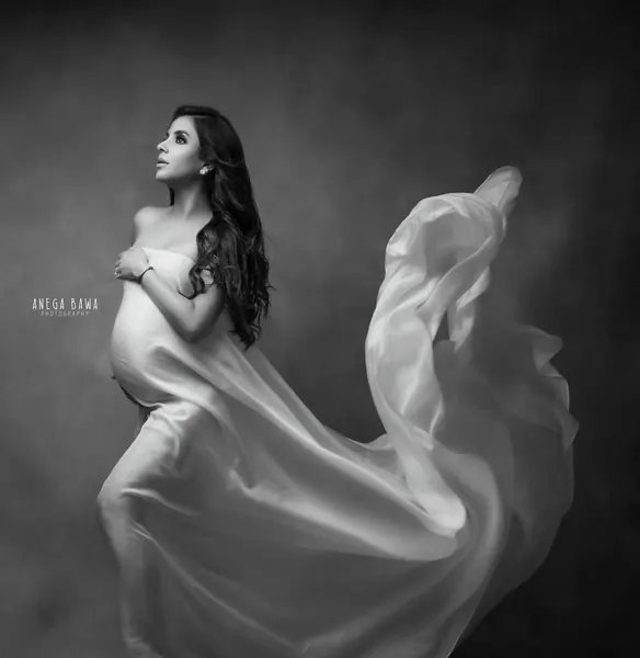 Black and white portrait of expecting mother in a sheer satin drape across her chest with fabric streaming behind her, looking up, hand on