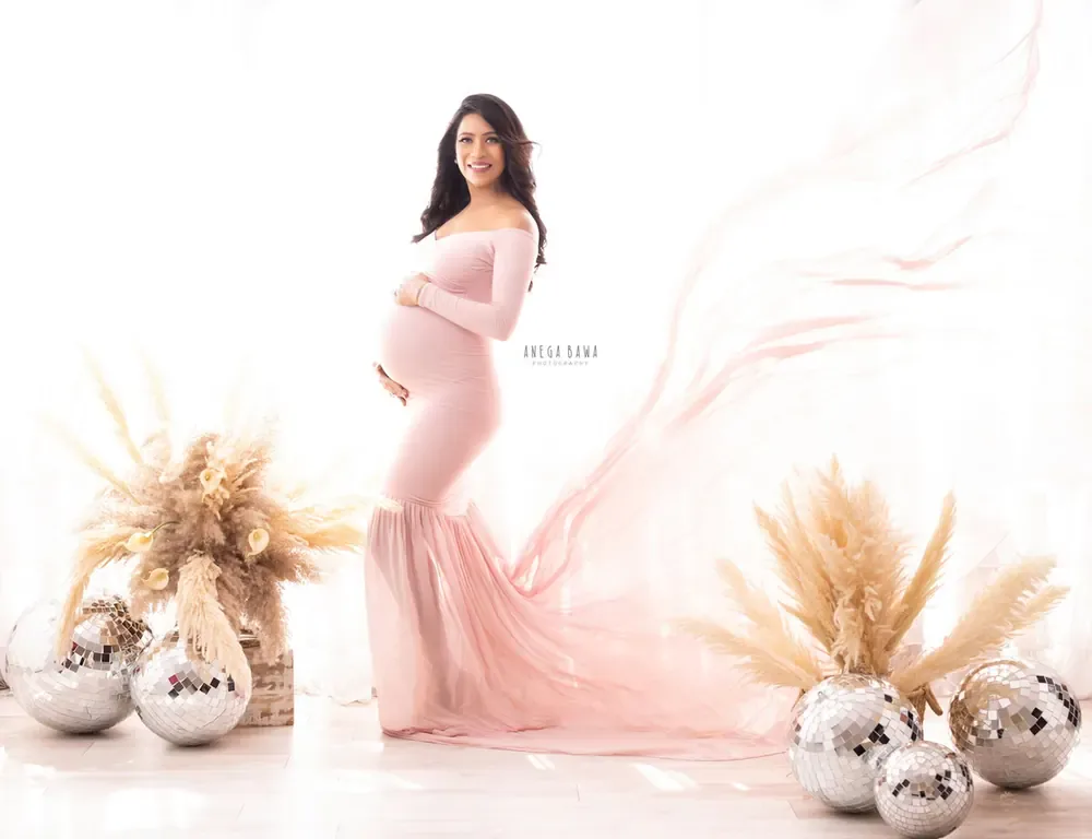 Expecting mother in a blush pink off-shoulder mermaid gown with a sheer pink train flying, surrounded by silver disco balls and pampas