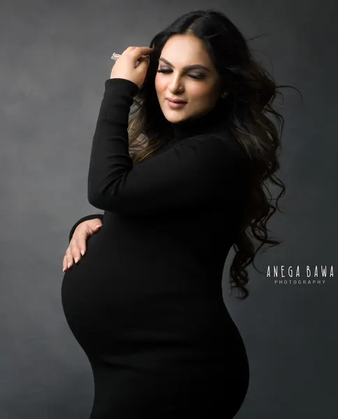 Expecting mother in a black turtleneck long-sleeve fitted dress, one hand in her wavy hair and the other on her bump, eyes closed, dark