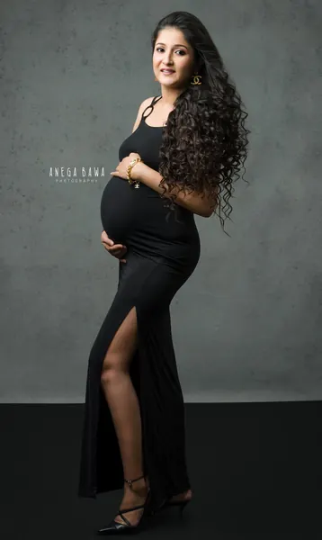 Expecting mother in a black strappy fitted gown with a high slit, black strappy heels and long curly hair, hands on her bump, grey backdrop.