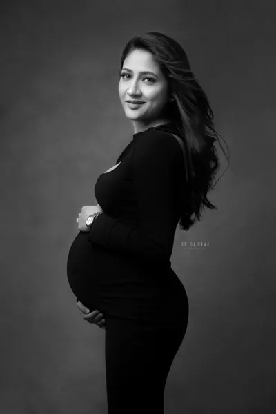 Black and white portrait of expecting mother in a fitted black long-sleeve dress looking over her shoulder with both hands under her bump