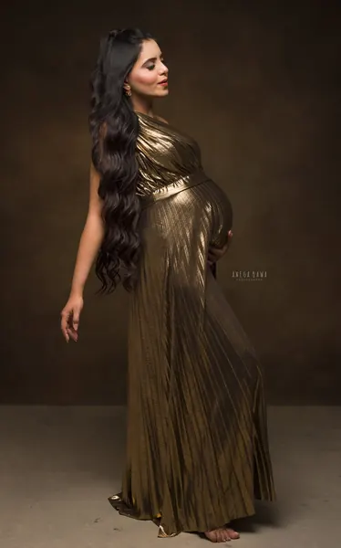 Expecting mother in a gold pleated metallic one-shoulder gown with a belt, very long wavy hair, barefoot, hand on her bump, looking to the