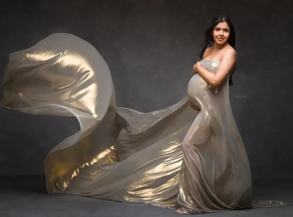 Expecting mother in a metallic champagne-gold strapless drape with a vast sheer metallic train sweeping out to the side, hands on her bump