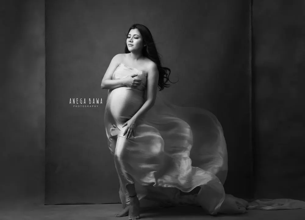 Black and white portrait of expecting mother kneeling in a sheer drape with fabric streaming behind her, bare bump, covering her chest with