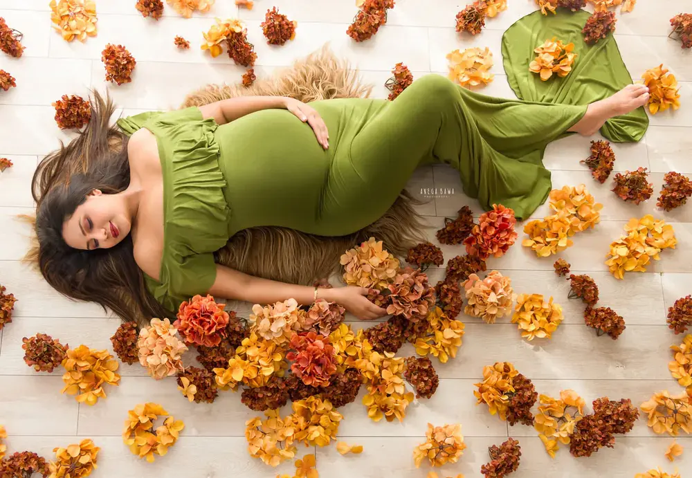 Overhead view of expecting mother lying on a fur rug in a green off-shoulder ruffle gown among orange and rust hydrangeas on a white wooden