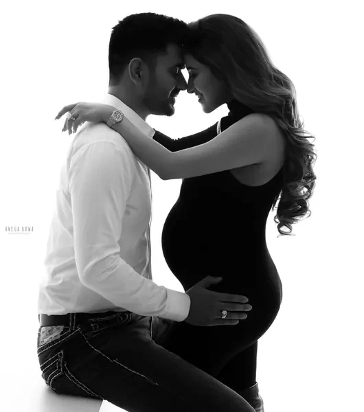 High-key silhouette of a couple seated face to face touching foreheads, husband in a white shirt and jeans with his hand on her bump, wife