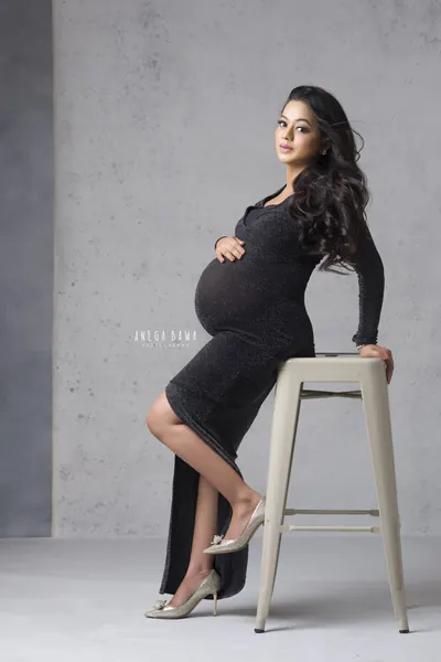 Expecting mother seated on a tall metal stool in a black glitter long-sleeve fitted dress with a slit and silver heels, long wavy hair