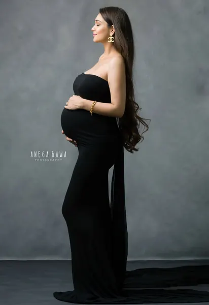 Expecting mother in profile in a black strapless fitted gown with a long draped train and gold bracelet, long hair, hand on her bump, grey