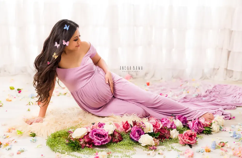Expecting mother reclining on a fur rug in a lilac off-shoulder gown with a long train, butterflies in her hair, surrounded by roses