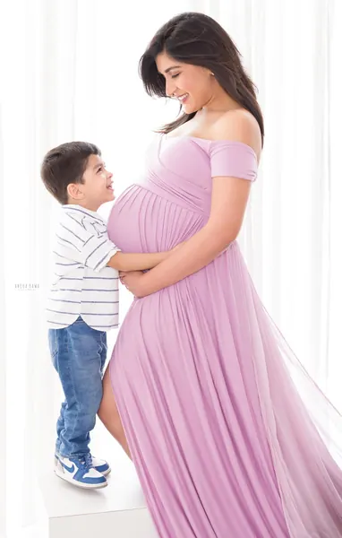 Young son in a striped shirt, jeans and sneakers hugging his smiling expecting mother, who wears a lilac pleated off-shoulder gown with a