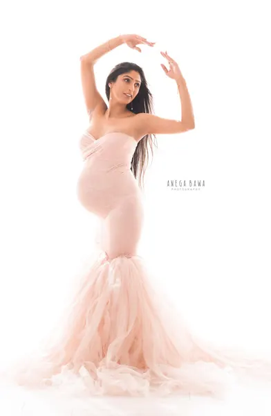 Expecting mother in a blush pink strapless mermaid gown with a layered tulle skirt, arms raised in a ballet pose, hand reaching above her