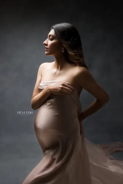 Expecting mother in a dusty rose satin drape clasped across her chest with bare shoulders, hair blowing in profile, hand on her hip, dark