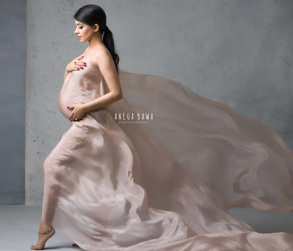 Expecting mother in a blush silk drape held to her chest with sheer fabric streaming behind her, walking, hair in a ponytail, hand on her