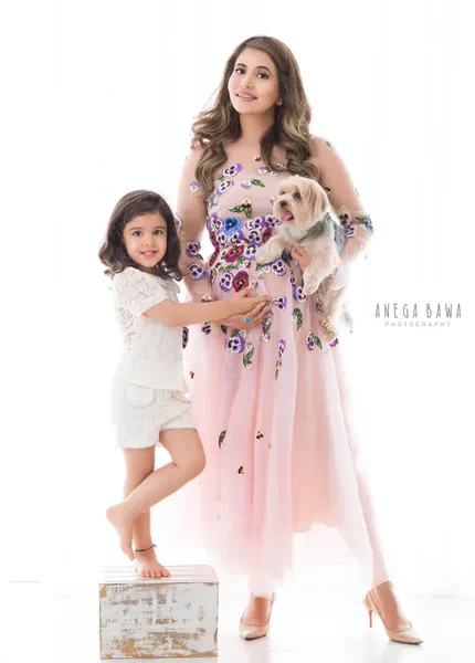 Family portrait: expecting mother in a pink tulle gown embroidered with pansy flowers and heels holding a small dog, her young daughter in