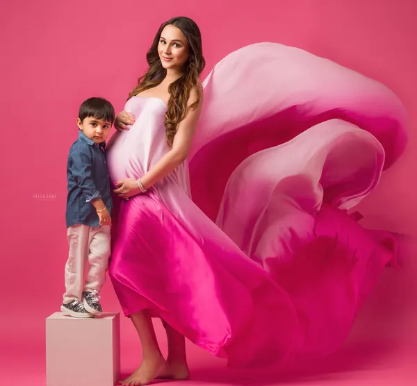Expecting mother in a pink ombre silk drape billowing out, standing with her young son in a denim shirt on a pink block, solid pink