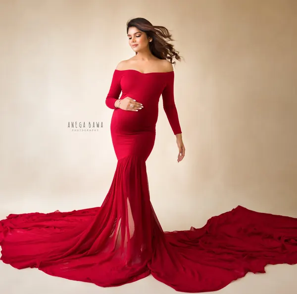 Expecting mother in a fitted red off-shoulder long-sleeve mermaid gown with a wide spread train on the floor, hair blowing, eyes down, warm